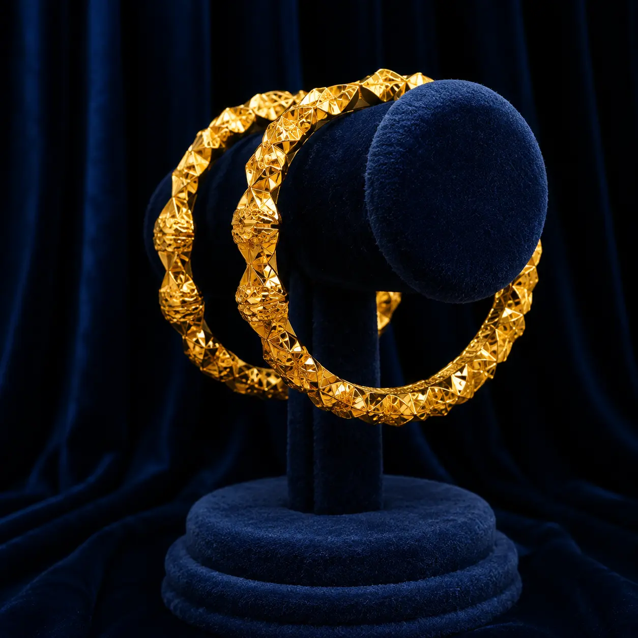 Pair of diamond-cut faceted gold bangles on a navy velvet stand